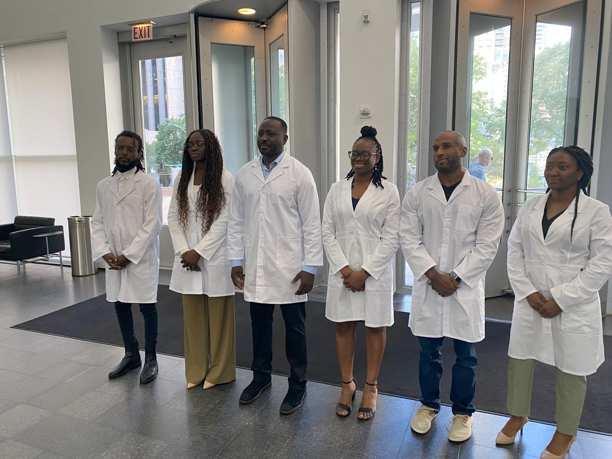 Six cast members in white lab coats standing in a row in a bright, modern atrium in Chicago